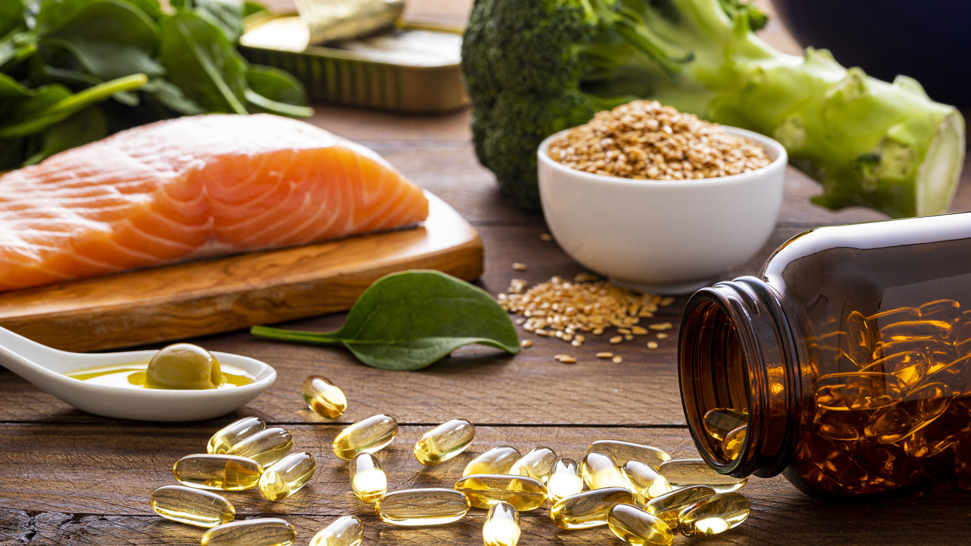 Front view of many fish oil capsules spilling out from the bottle surrounded by an assortment of food rich in omega-3 such as salmon, flax seeds, broccoli, sardines, spinach, olives and olive oil.